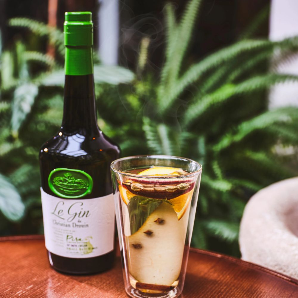 Quel gin choisir ? Le gin de Christian Drouin Pira accompagné d’un cocktail à base de poire, d’orange et d’épices, servi sur une table en bois avec plantes vertes en arrière-plan. Le Club des Connaisseurs