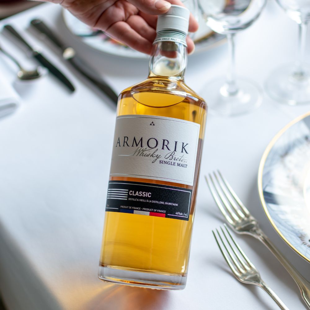 Bouteille de whisky Armorik Classic tenue à la main au-dessus d'une table dressée, avec des couverts et des verres en arrière-plan. Le Club des Connaisseurs.