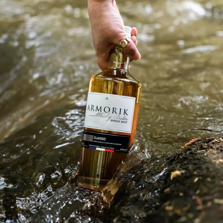 Bouteille de whisky Armorik Classic immergée dans une rivière, tenue par une main ornée d'une bague. Le Club des Connaisseurs.