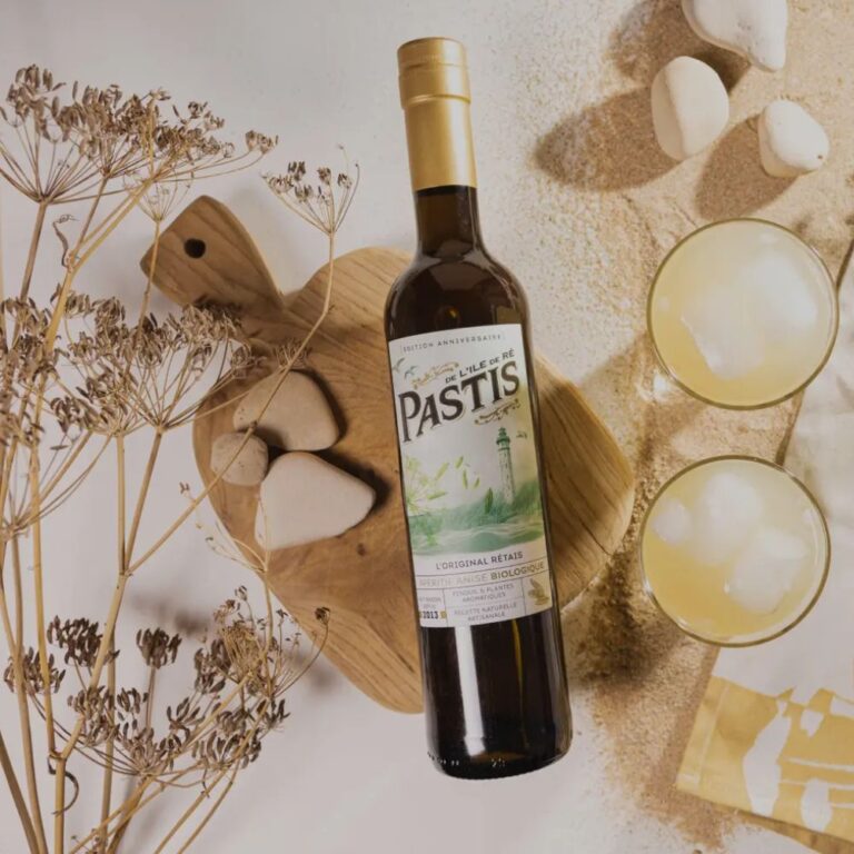 Bouteille de Pastis de l’Île de Ré, apéritif anisé biologique, posée sur une planche en bois avec des plantes aromatiques séchées, des galets et deux verres servis sur fond sableux. Pastis artisanal français élaboré sur l’Île de Ré.