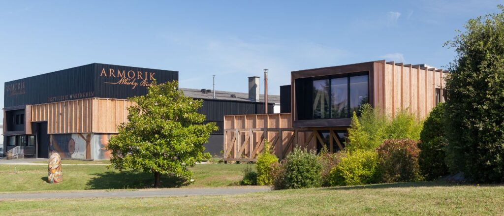 Bâtiment moderne et traditionnel de la distillerie Armorik sous un ciel bleu. Façade noire et bois avec le logo Armorik Whisky Breton. Espaces verts et arbustes autour des installations. Disponible sur Le Club des Connaisseurs.