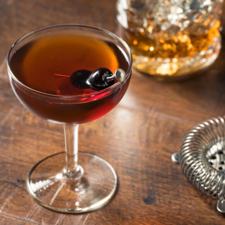 Cocktail Manhattan servi dans un verre à pied avec une cerise en garniture, accompagné d’un verre de whisky en arrière-plan. Un cocktail classique à base de whisky, vermouth rouge et bitters. Le Club des Connaisseurs.