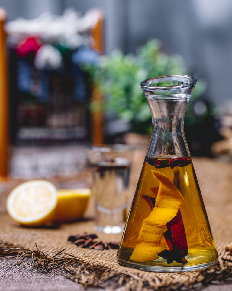 Carafe en verre contenant du rhum arrangé avec des zestes d’orange, un bâton de cannelle et des étoiles de badiane, posée sur une table en toile de jute avec un demi-citron en arrière-plan. Le Club des Connaisseurs.