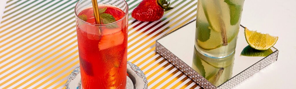 Bannière article de blog sur le mojito fraise. Cocktail mojito fraise rafraîchissant servi dans un verre à cocktail sur une table rayée, avec une fraise et une tranche de citron vert en décoration. Le Club des Connaisseurs.