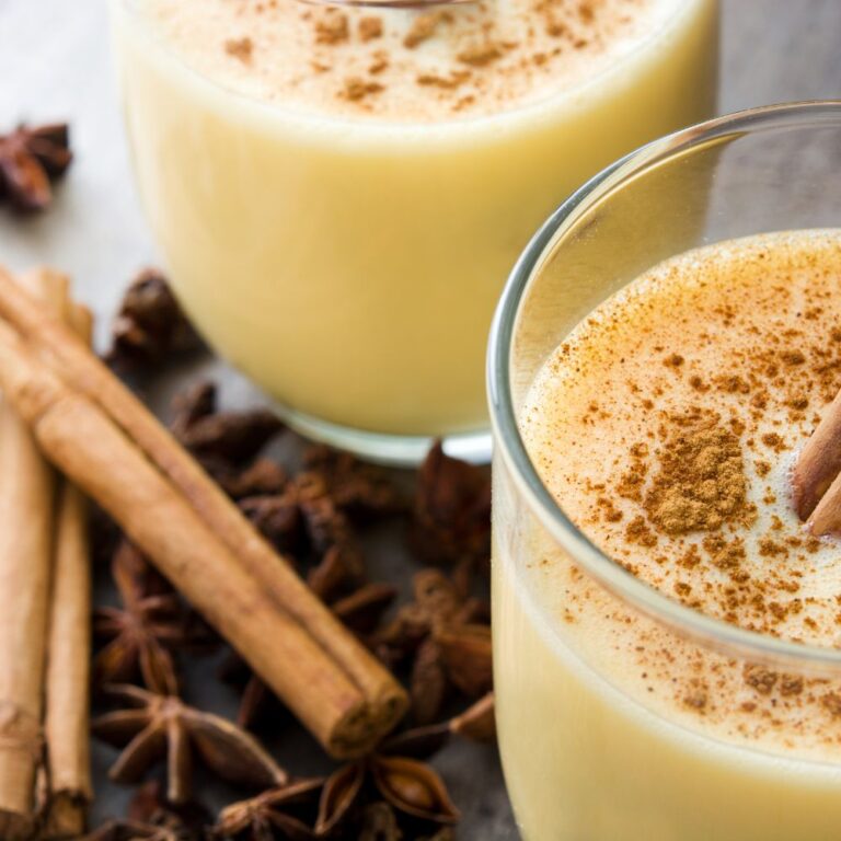 Miniature article de blog Eggnog Le Club des Connaisseurs. Gros plan sur un verre de lait de poule saupoudré de cannelle, entouré d’épices comme la cannelle. cocktail crémeux hivernal.