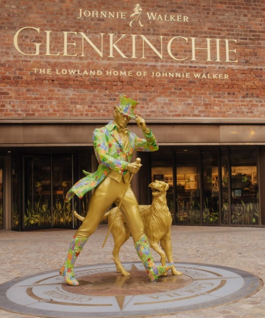 Image article de blog La route du whisky en écosse Le club des connaisseurs : Statue colorée du marcheur iconique devant la distillerie Glenkinchie en Écosse, maison du whisky Johnnie Walker.