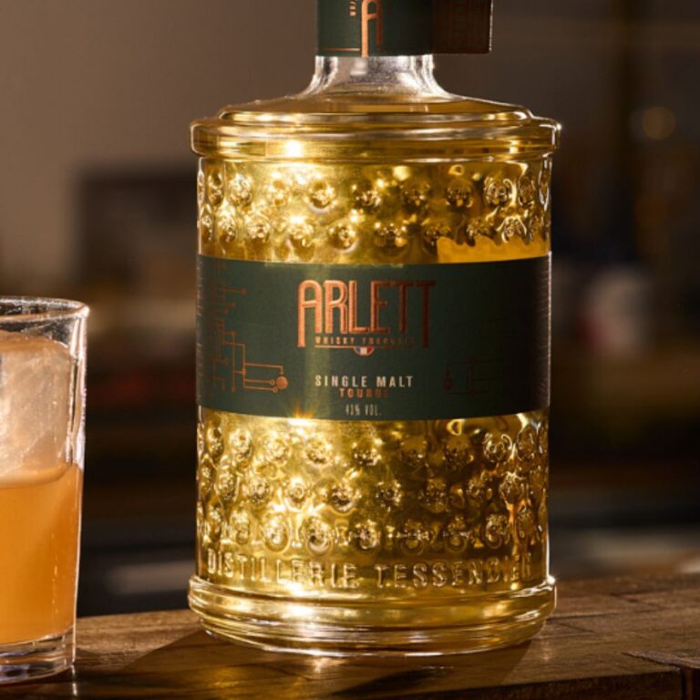 Bouteille de whisky français Arlett Single Malt Tourbé de la Distillerie Tessendier, mise en avant sur un bar avec verre à whisky en ambiance chaleureuse, photo packshot pour page marque Arlett sur le Club des Connaisseurs.
