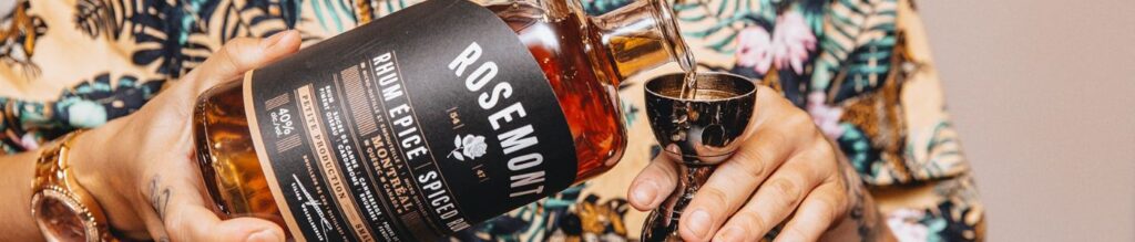 Image page marque Rosemont distillerie de Montréal : Service de rhum épicé Rosemont dans un jigger, gros plan sur l’étiquette noire et mains tatouées en arrière-plan tropical.