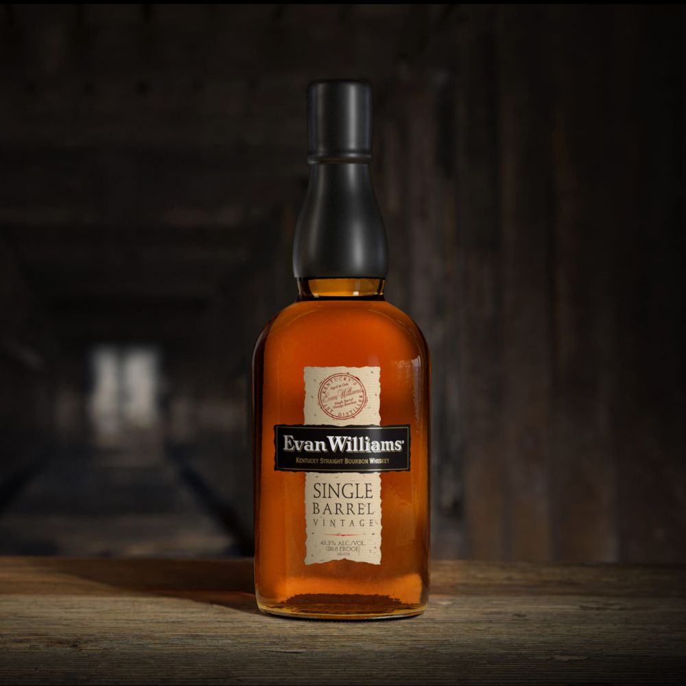Beautyshot de la bouteille de bourbon Evan Williams Single Barrel Vintage posée sur une table en bois, avec un fond sombre évoquant un chai.