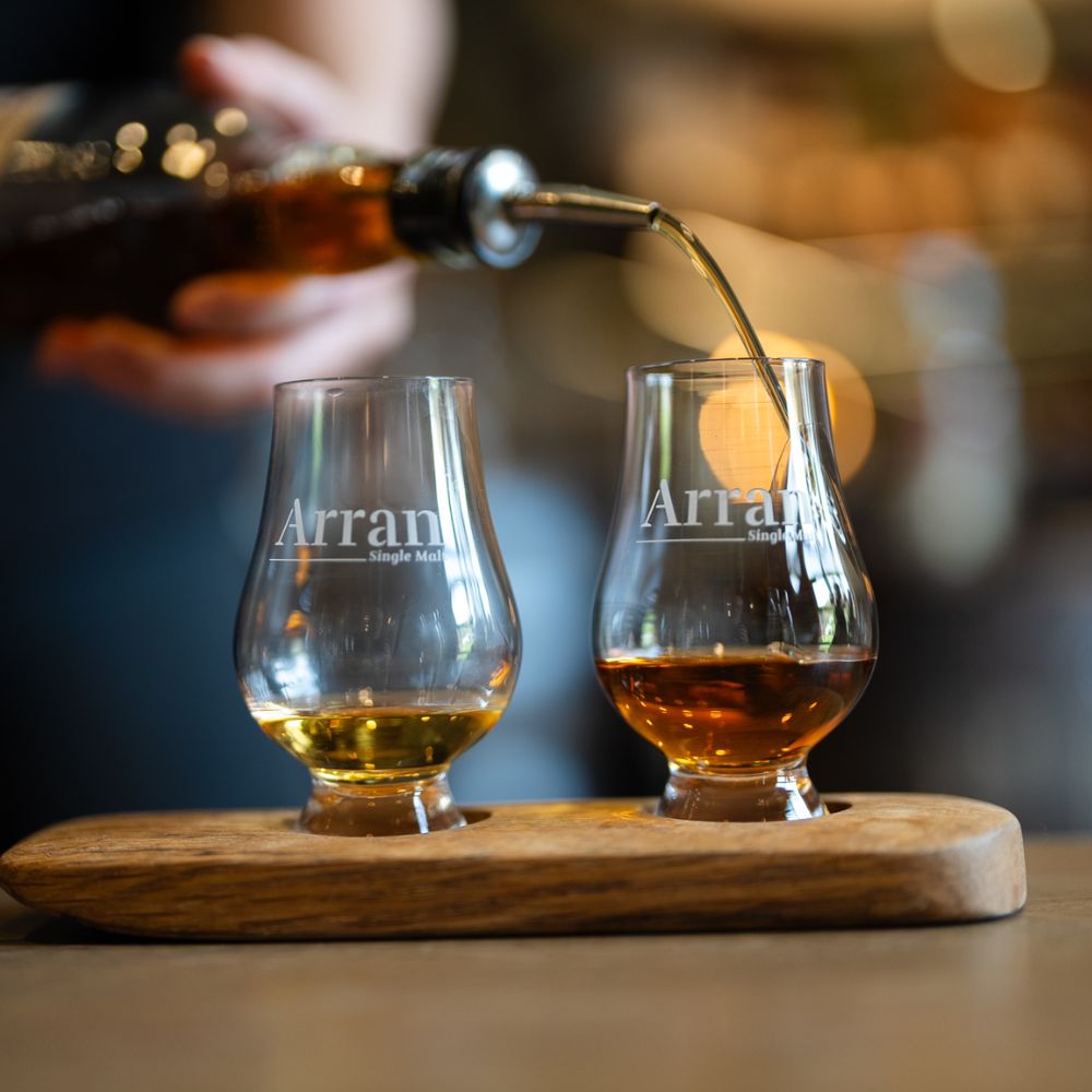 Dégustation de whisky Arran Single Malt avec deux verres tulipe sur plateau en bois, versement en cours, ambiance chaleureuse de distillerie.