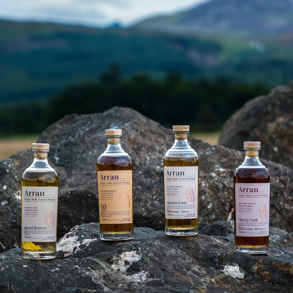 Bouteilles de whisky Arran Single Malt posées sur des rochers en Écosse avec vue sur les montagnes, gamme complète Barrel Reserve, 10 ans, Quarter Cask et Sherry Cask.